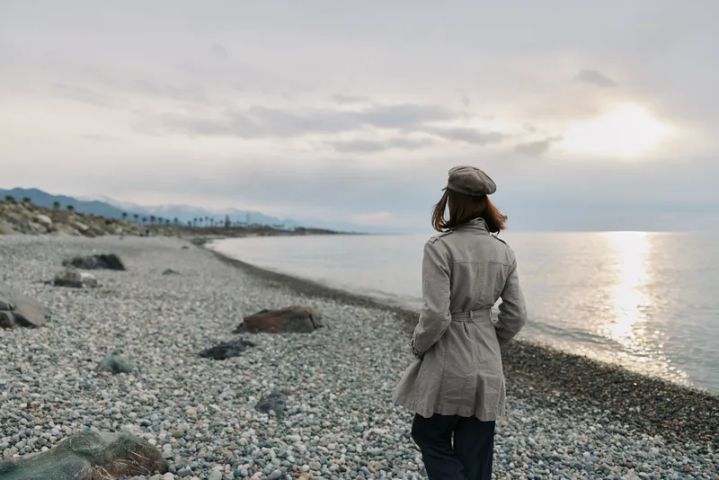 A woman wearing a coat and hat walks alone on a pebble beach during cloudy sunset. The calm sea and distant mountains create a peaceful and reflective mood.