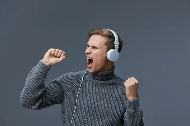 Vibrant image of a man enjoying music with headphones, expressing joy and excitement. He is wearing a cozy gray sweater, raising his fists, and showing enthusiasm against a plain solid background. Con