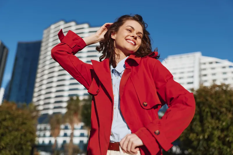 Young woman in a red trench coat smiles joyfully while adjusting her hair outdoors on a sunny day with modern buildings in the background.