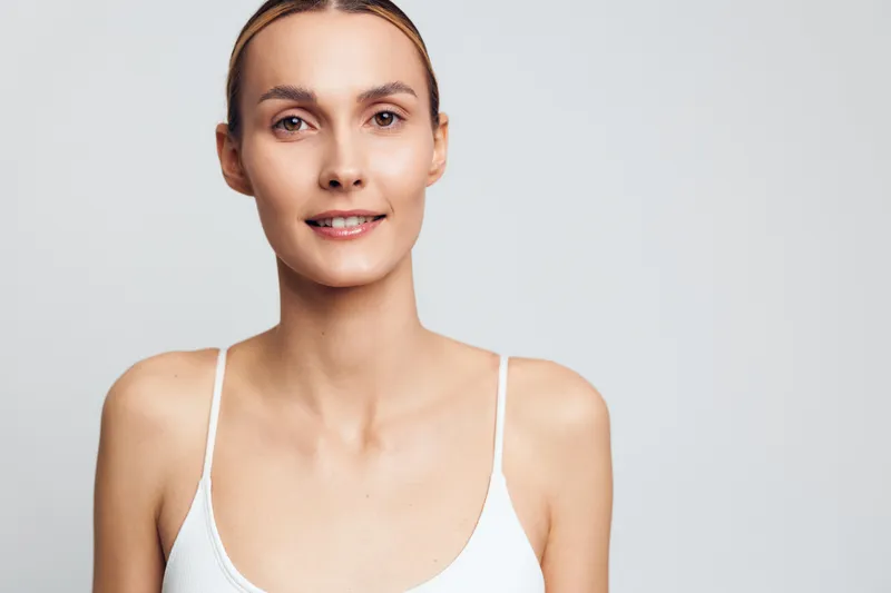 Young woman with blonde hair and clear skin smiles gently while wearing a white tank top against a plain light background in a natural studio portrait.