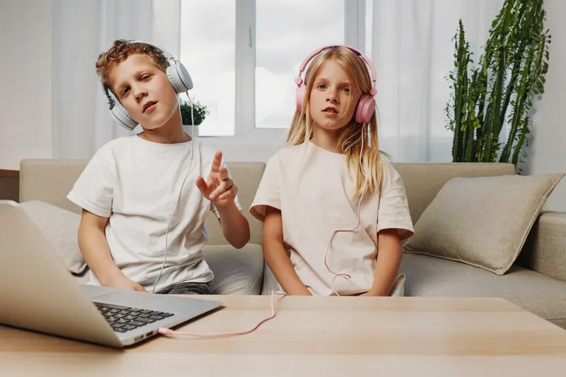 Two children wearing headphones listen to music together at home, sitting on a sofa with a laptop on the table. The scene conveys childhood leisure, technology, and cozy indoor activity.