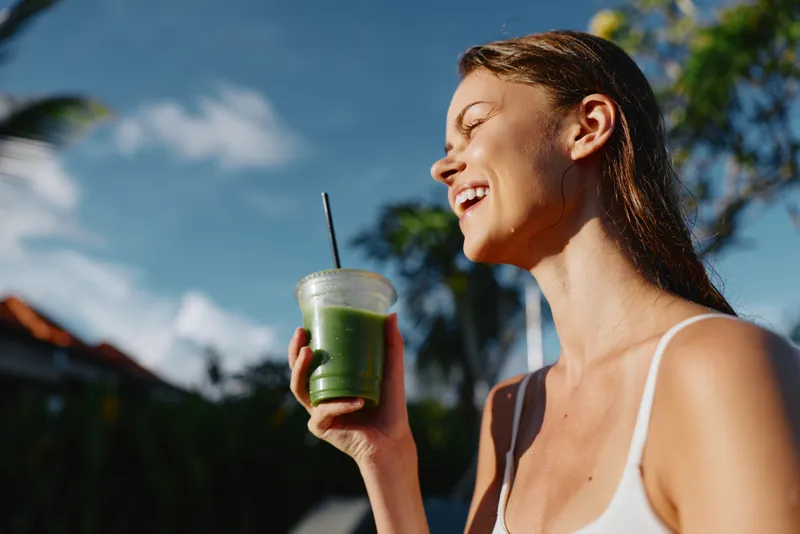 Smiling woman enjoying a green smoothie outdoors on a sunny day, casual summer vibe. Close-up of the drink and cup, straw, fresh ingredients, relaxed wellness mood.