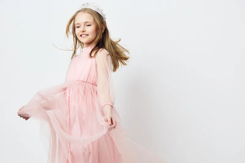 A cheerful girl in a flowing pink dress twirls joyfully against a soft white background, embodying innocence and playfulness