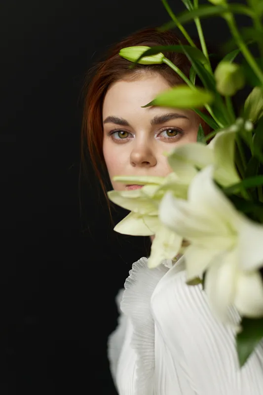 Young model with flowers, soft natural makeup in white clothing, showcasing beauty and serenity against a dark background, perfect for beauty, lifestyle, and floral themes