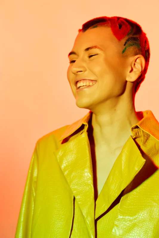 Smiling woman with bright red hair and a vibrant yellow jacket, radiating happiness and confidence against a warm colorful background, showing modern style and joy.