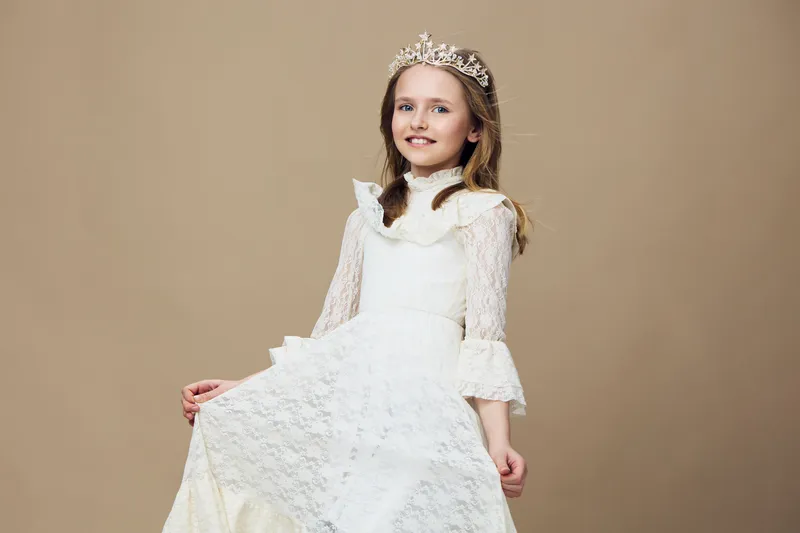 Young girl in an elegant white dress with a crown, smiling against a beige background, perfect for children s fashion and special occasions