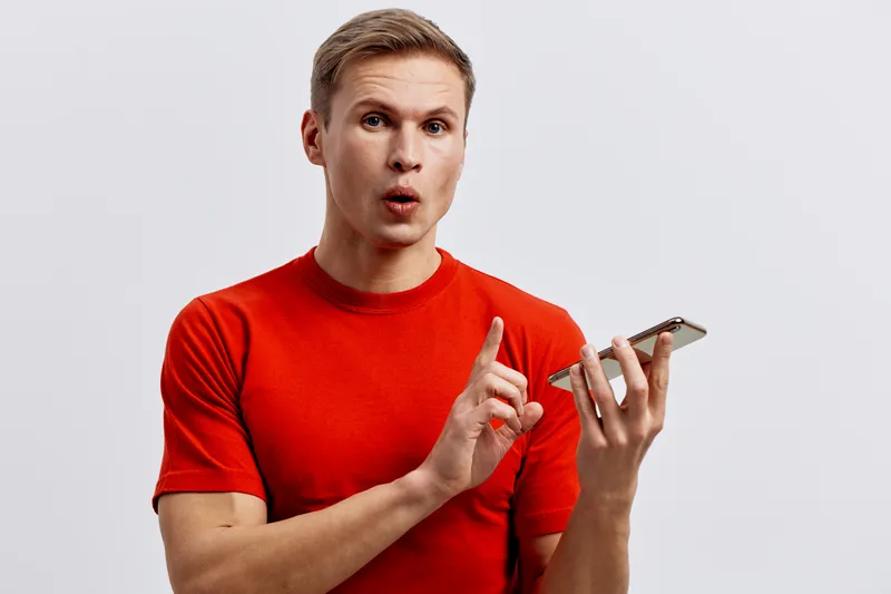 Young man with surprised facial expression, wearing red t-shirt, holding smartphone, isolated on plain background. Model reacting with curiosity or shock during mobile usage, studio portrait. People l