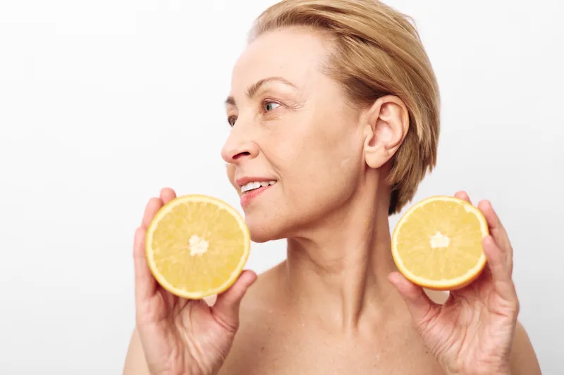 Mature woman with short hair holds two orange halves near her face, smiling slightly against a white background. Concept of natural skincare, healthy lifestyle, and fresh fruit benefits.