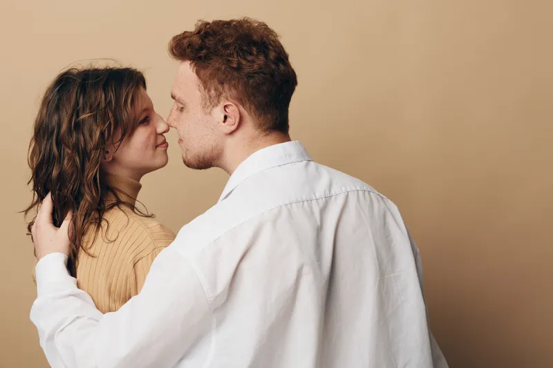 Young couple embraces and kisses gently on beige background. Intimate moment shows love, affection, and connection between man and woman in casual clothing.