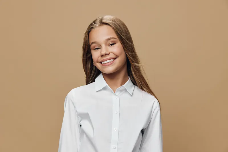 Girl, smiling and happy with long hair wearing white shirt standing against beige background. Child, confident and positive with casual clothing for lifestyle portrait indoors.