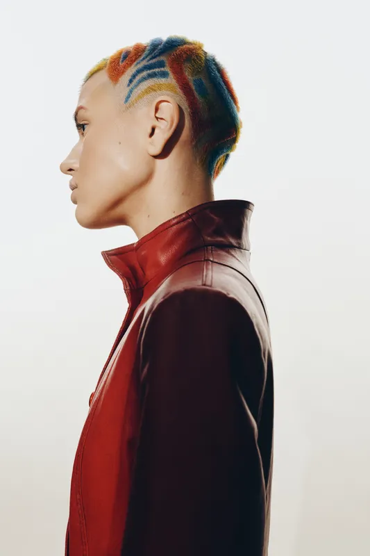 Profile of a woman with colorful braided hair styled in a modern pattern, wearing a red jacket, with a minimalist bright background and sharp contrast.