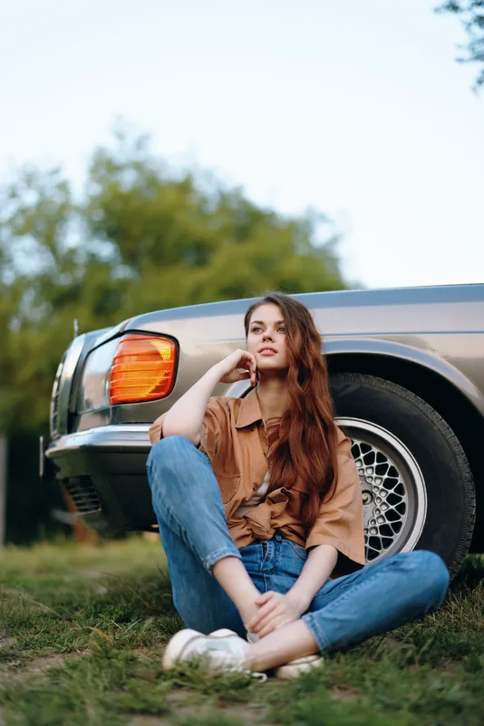 Young woman with long hair sitting on the grass next to a vintage car, wearing casual clothes, showcasing a relaxed and thoughtful expression in the park.