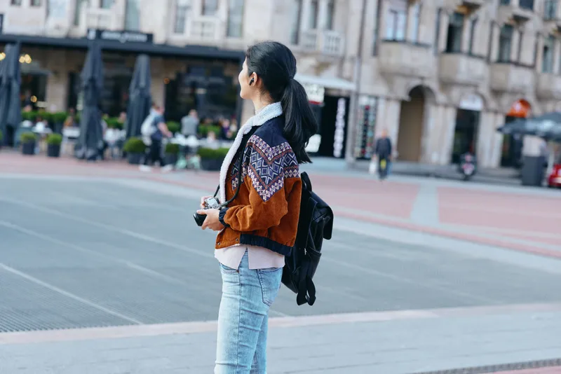 Woman, street, backpack, urban, casual, fashion, jacket, jeans, camera, travel on a bright plaza capturing urban style, exploration and contemporary outerwear in a busy city