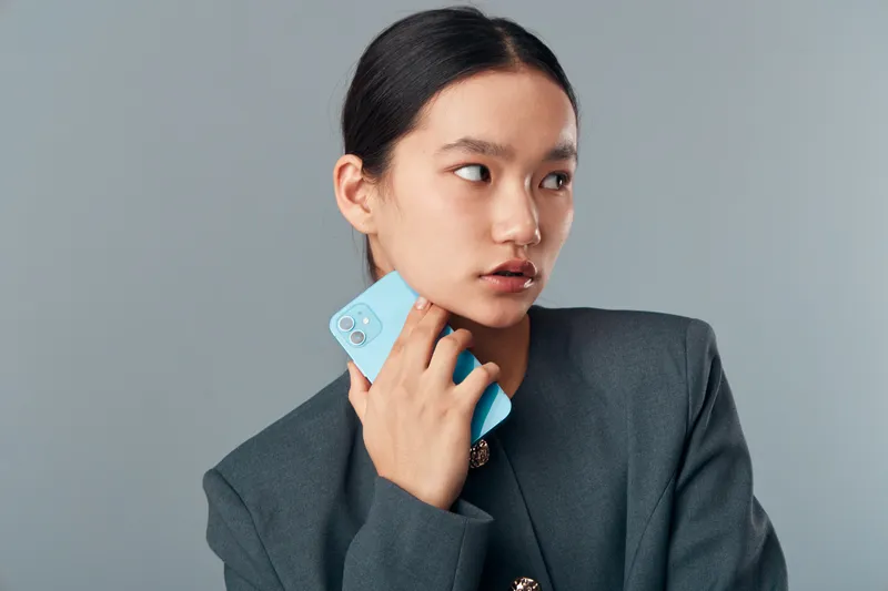 Young woman in gray blazer holding light blue smartphone near face looking to side on gray background. Portrait of professional female with modern device in minimal studio for communication or