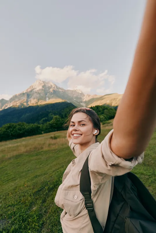 Woman lifestyle portrait outdoors in the mountains, smiling selfie moment, wearing backpack, enjoying nature on a clear day, vibrant landscape and open sky