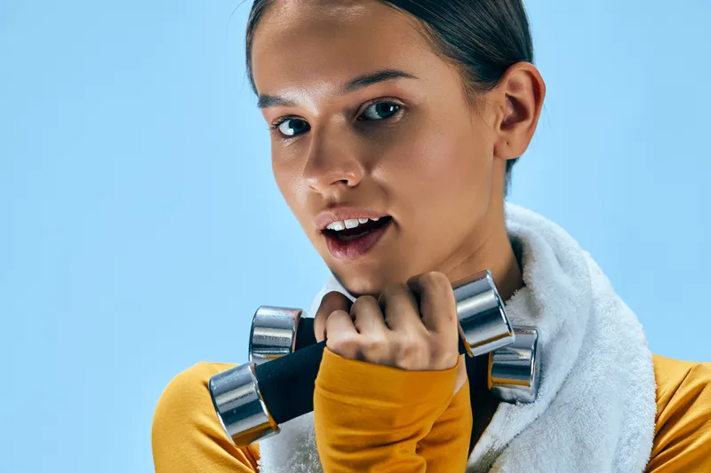 Woman fitness athlete lifts dumbbells with a towel around her neck, intense focus and energy in a bright studio, capturing determination during a workout.