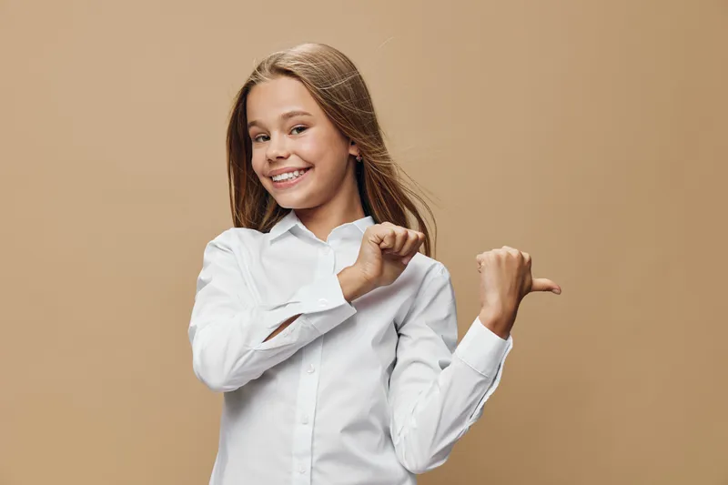 Girl in white shirt smiling and pointing with thumb to side on beige background. Child with long hair wearing casual clothing showing gesture and looking at camera with happy expression.