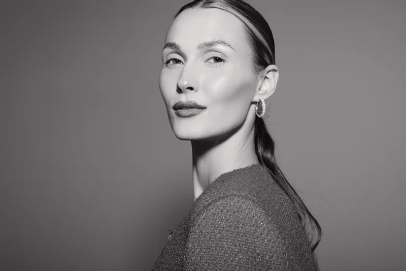 Portrait of a fair skinned Caucasian woman with slicked back hair wearing hoop earrings and a textured top. Studio lighting highlights her confident expression and clear skin.