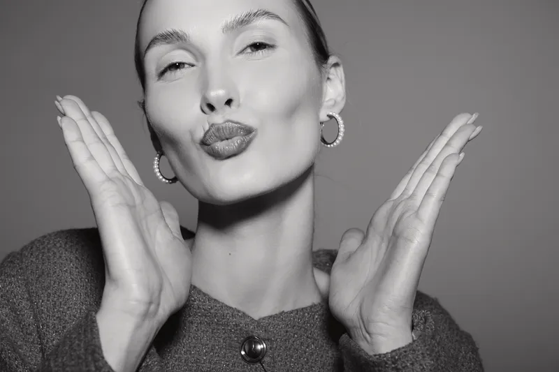 Young Caucasian woman with fair skin blowing a kiss, smiling with closed eyes, wearing hoop earrings and a buttoned sweater on a grey background. Concept of affection and beauty.
