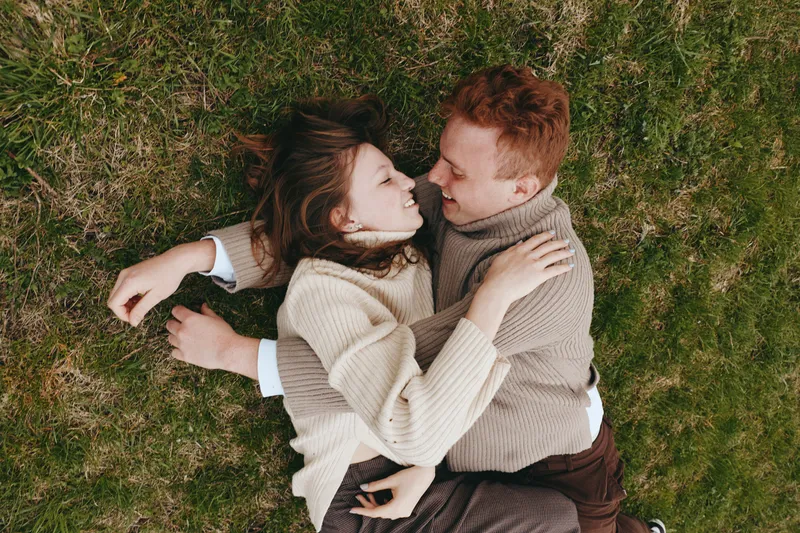 A couple lying on grass shares an intimate moment, embracing in cozy knitwear under soft daylight. A tender scene of connection, affection, and outdoor lifestyle photography. Perfectly candid