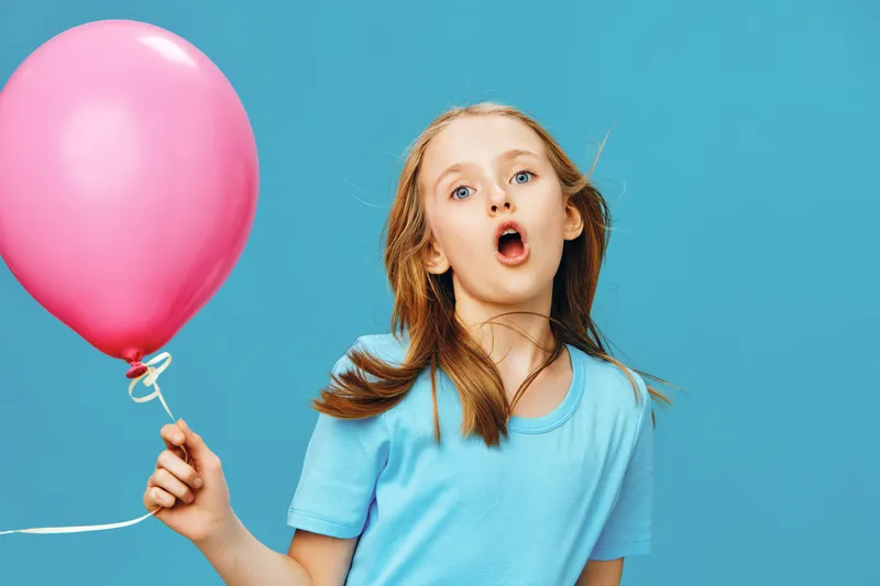 Young girl with a surprised expression holding a pink balloon against a blue background, showcasing joy and playfulness in a vibrant setting