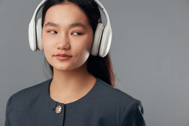 Young woman wearing white headphones and dark jacket looks aside on gray background. Portrait of confident girl with long hair listening to music or audio in casual style.