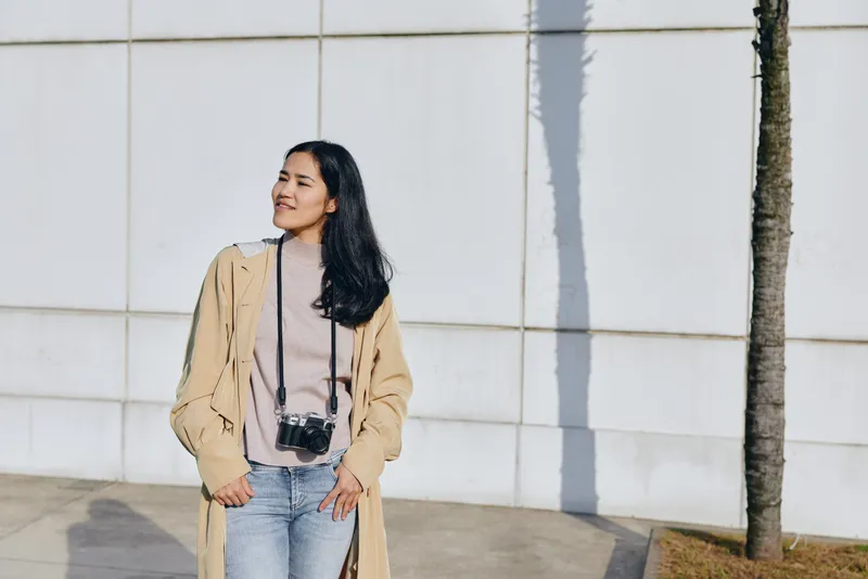 Woman wearing a long beige coat and jeans with a camera, standing beside a white wall in an urban street setting, capturing candid fashion and travel vibes