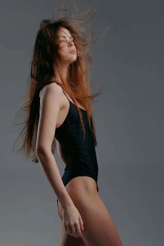 Young woman in black swimsuit with windblown hair posing against grey background