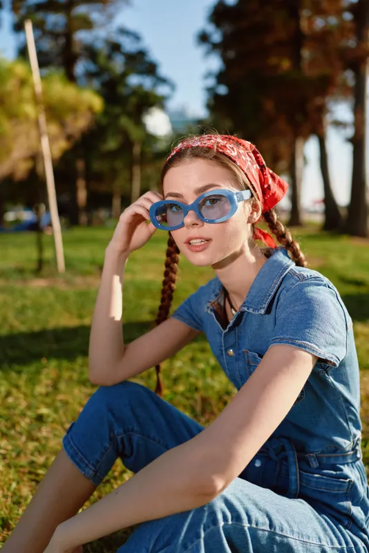 lifestyle woman wearing modern boho-western denim outfit with blue sunglasses sitting outdoors in sunlight in film color street style mood