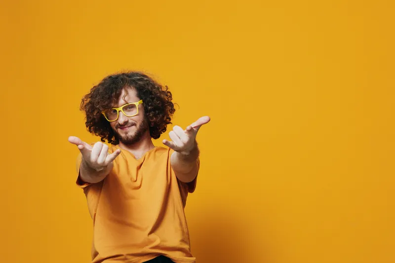 Cheerful young man with curly hair in yellow glasses on a vibrant orange background, expressing joy and positivity while wearing a casual shirt