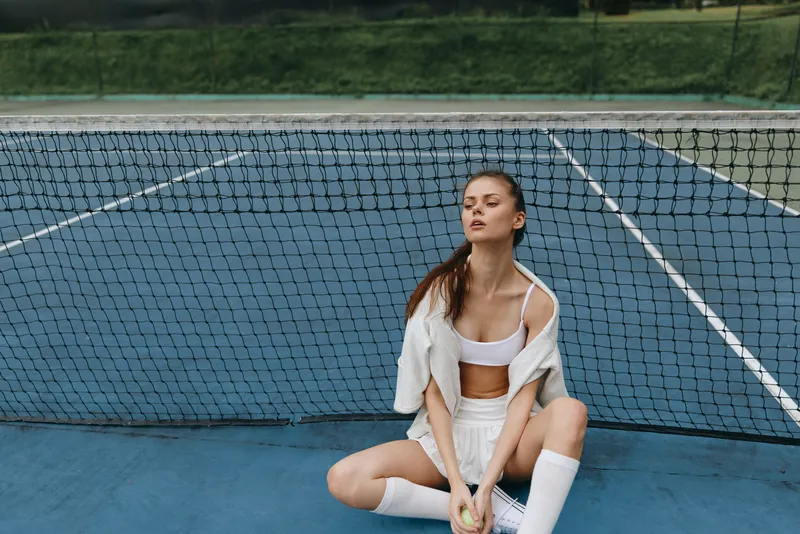Young female tennis player in white sportswear, sitting on the court with a tennis ball, showcasing determination and focus, on a sunny day