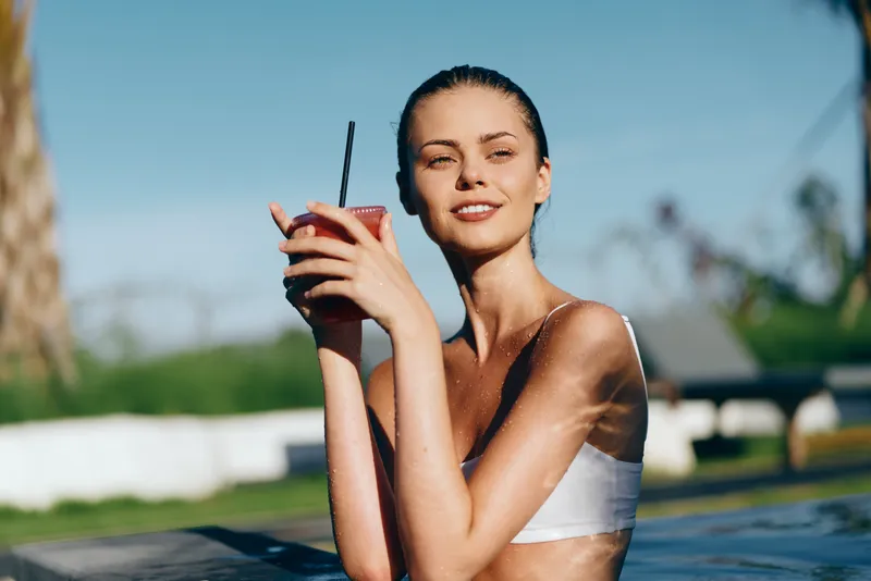 Young woman in white bikini holding a drink with a straw relaxing in the pool on a sunny day with clear blue sky and palm tree in the background.