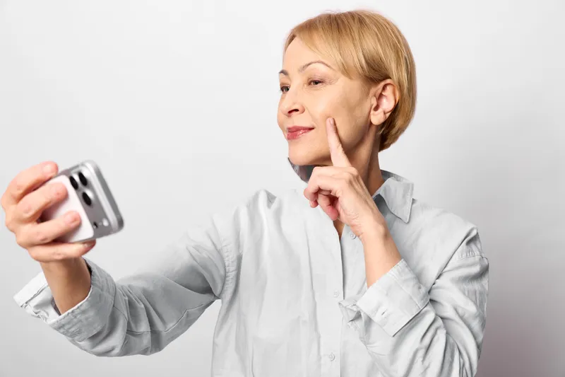 Woman takes selfie with smartphone in studio, white background, thinking expression, hand gesture near cheek, holding phone at arm length, casual shirt, clean portrait for social media