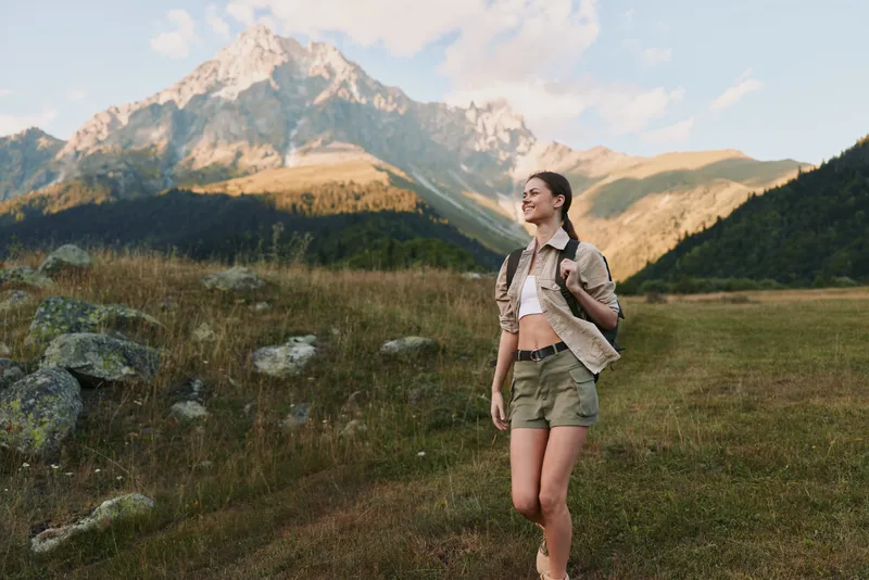 Young woman explores a mountain meadow in casual hiking attire with a backpack, bathed in golden hour light as distant peaks loom, conveying outdoor adventure.