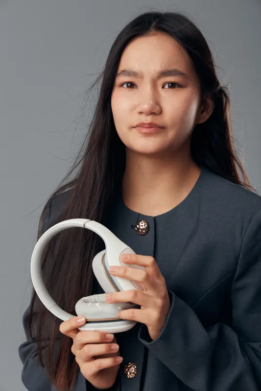 Young woman holding white headphones with serious face, wearing dark jacket, studio portrait on gray background, technology accessory and lifestyle concept in closeup.