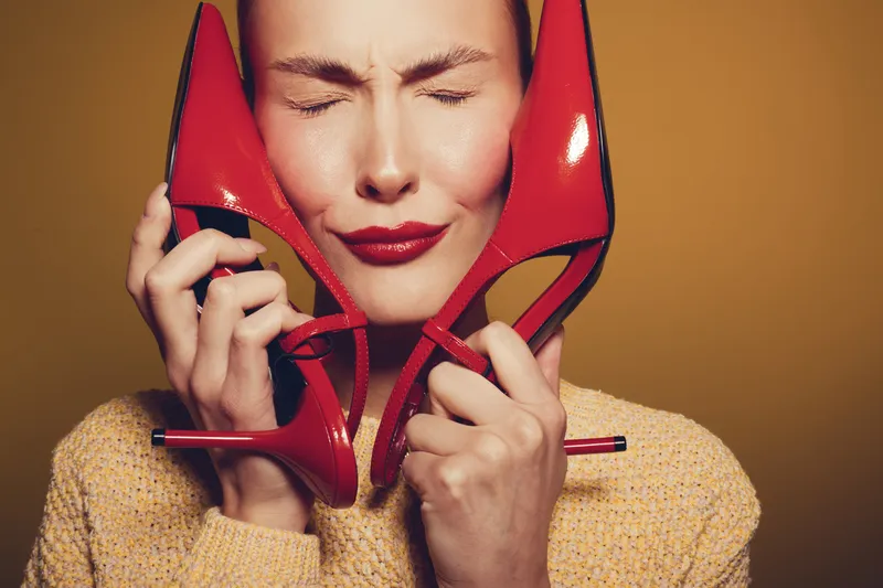 Young woman with closed eyes holds red high heels near face, wearing yellow sweater. Concept of fashion, style, and emotional expression on warm brown background.