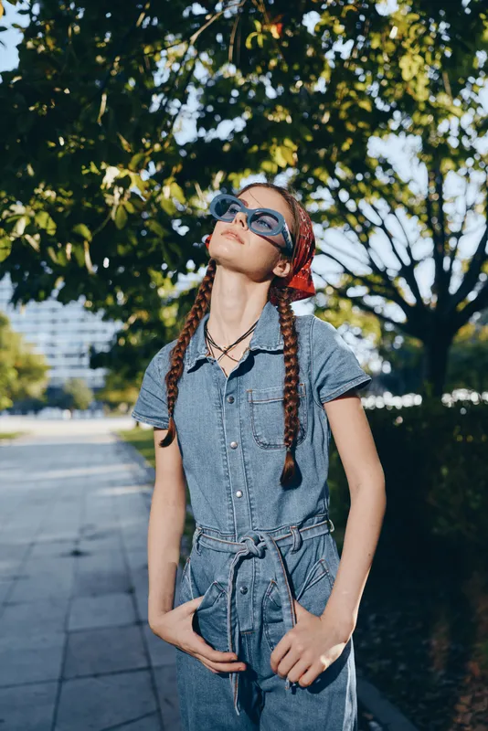 young woman with braids wearing sunglasses and denim outfit enjoying sunny day outdoors in urban park surrounded by green trees and modern buildings