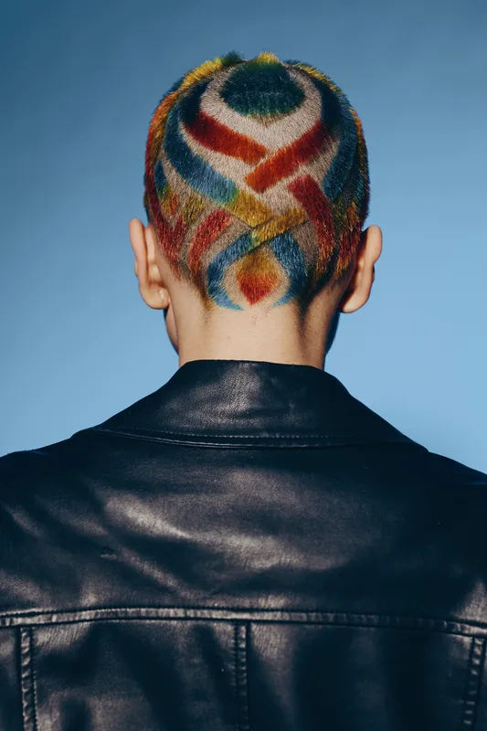 Woman with colorful dyed short hair patterns wearing a black leather jacket against blue background, showcasing creative style and bold fashion expression.