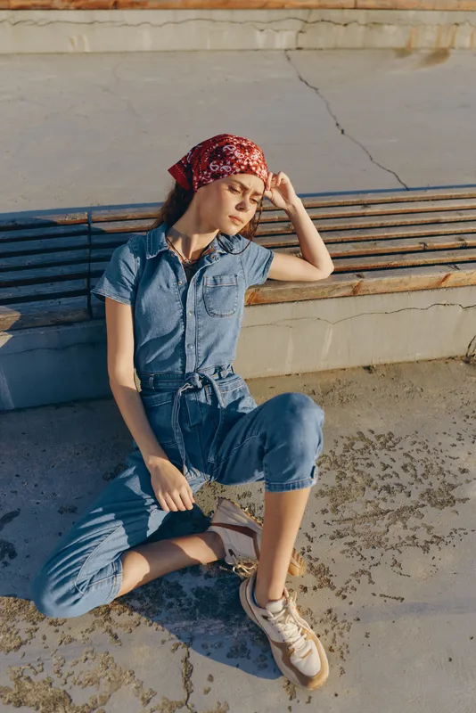 Lifestyle woman in modern boho-western denim street style sits relaxed outdoors wearing a red bandana with film color tones enhancing a vintage casual vibe.