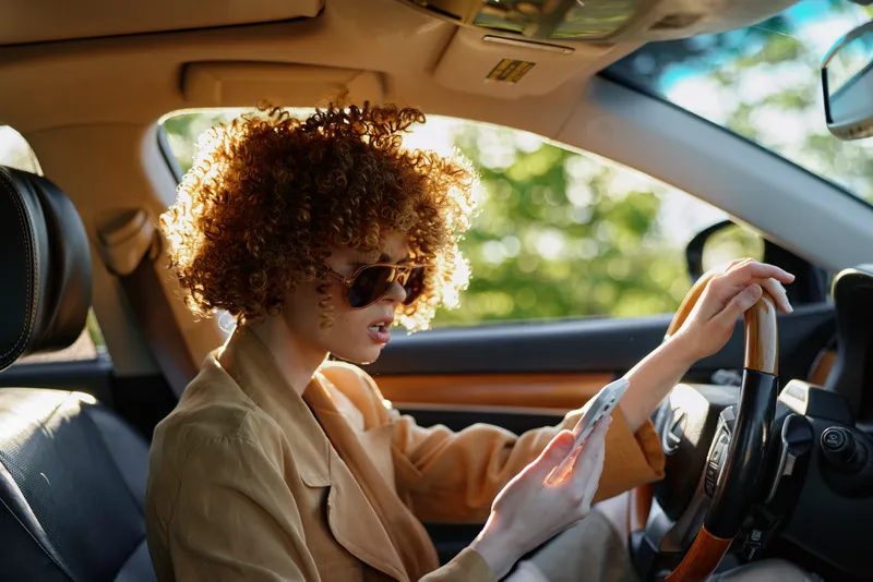 Stylish woman with curly hair driving a car, wearing sunglasses and a light blazer, captured in warm retro film tones, conveying a casual, confident lifestyle.