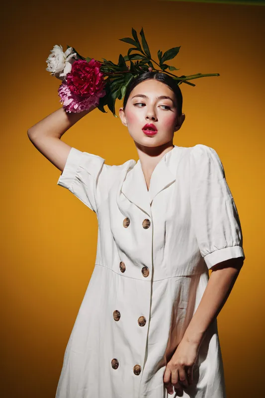 Stylish woman in a white dress wears a creative floral headpiece with vibrant red and white flowers. Bold makeup highlights fashion and beauty against yellow background.
