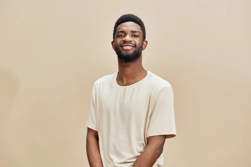 Smiling young Black man wearing a light beige t shirt, standing against a neutral toned background, exuding confidence and warmth in a modern style