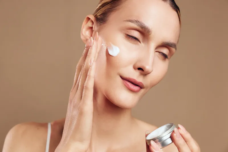Young woman applies moisturizing cream on face with hand holding jar, skincare routine concept, natural beauty, smooth skin, neutral background, closeup portrait.