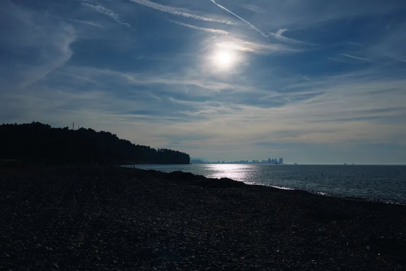 Pebble beach at dusk with calm sea, silhouetted shore and shimmering water under a clear sky. Soft light reflects on waves, creating a tranquil coastal scene with a distant horizon.