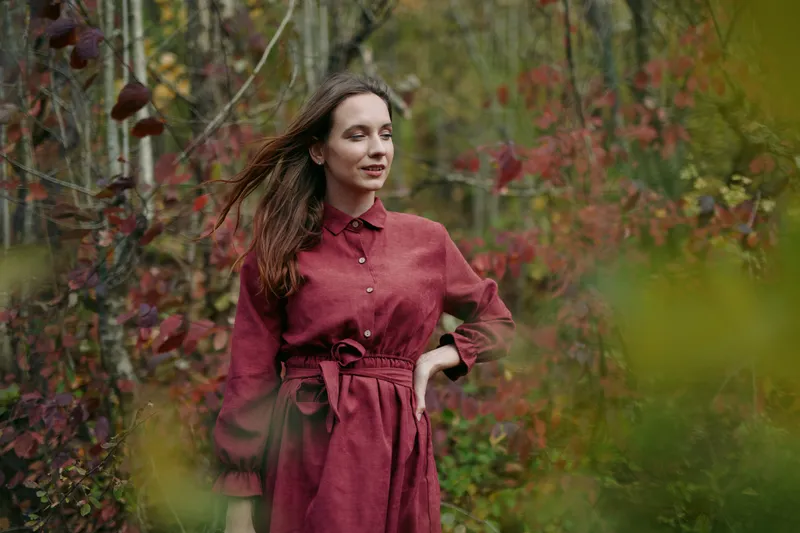 A candid portrait of a woman in a rustic red dress standing among autumn foliage, conveying genuine authenticity and quiet confidence with natural light and a truthful expression.