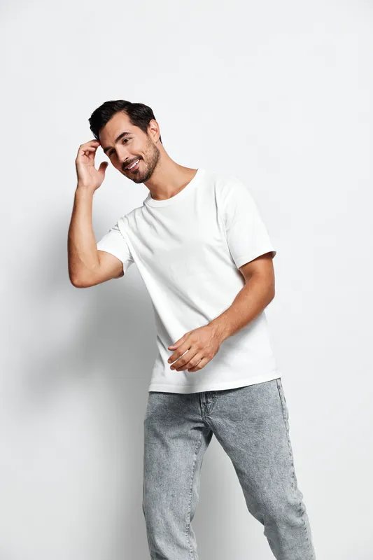 Smiling man dressed in casual white t-shirt and grey pants looking at the camera, showing friendly gesture while standing against a plain light background.