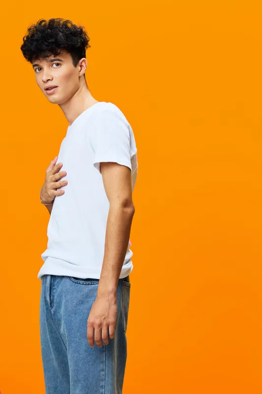 Young man standing against bright orange background wearing casual white t-shirt and blue jeans with thoughtful expression and curly dark hair.
