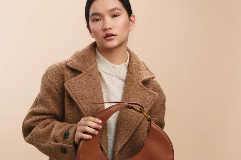 Asian woman wearing a warm tan coat and light sweater stands with a brown handbag in a neutral studio. She looks at the camera with a calm smile, radiating polished style and everyday chic.