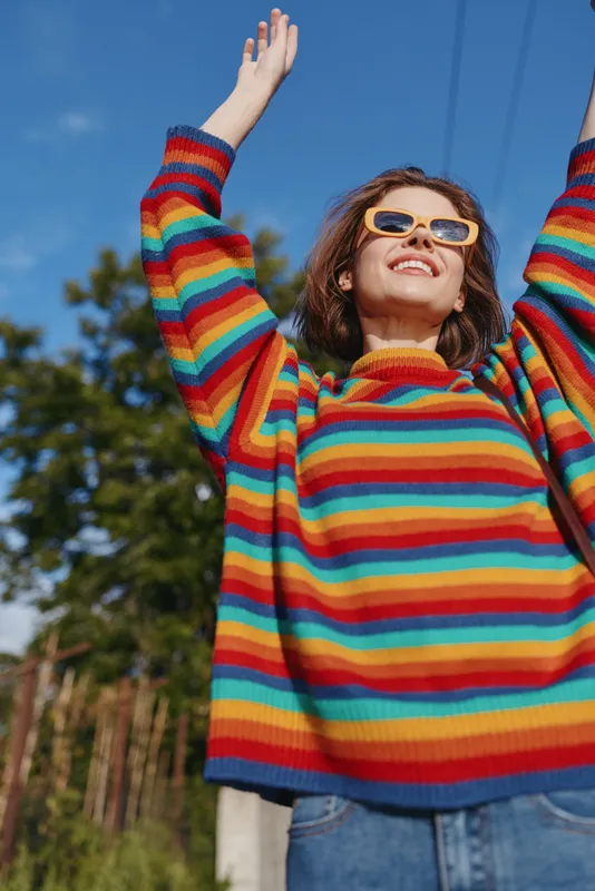 woman in a rainbow sweater smiling outdoors with sunglasses and arms raised, young adult posing in nature under sunny blue sky, casual outfit and joyful confident mood for lifestyle