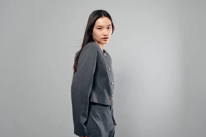 Young woman posing with gray suit and long dark hair standing against plain background in studio for fashion or portrait photography with confident expression.